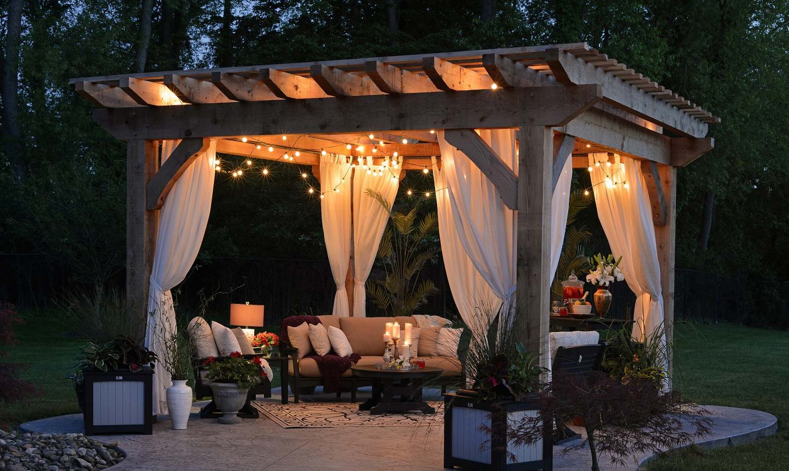 Osvětlená dřevěná pergola s bílými závěsy a pohodlným posezením večer na zahradě.